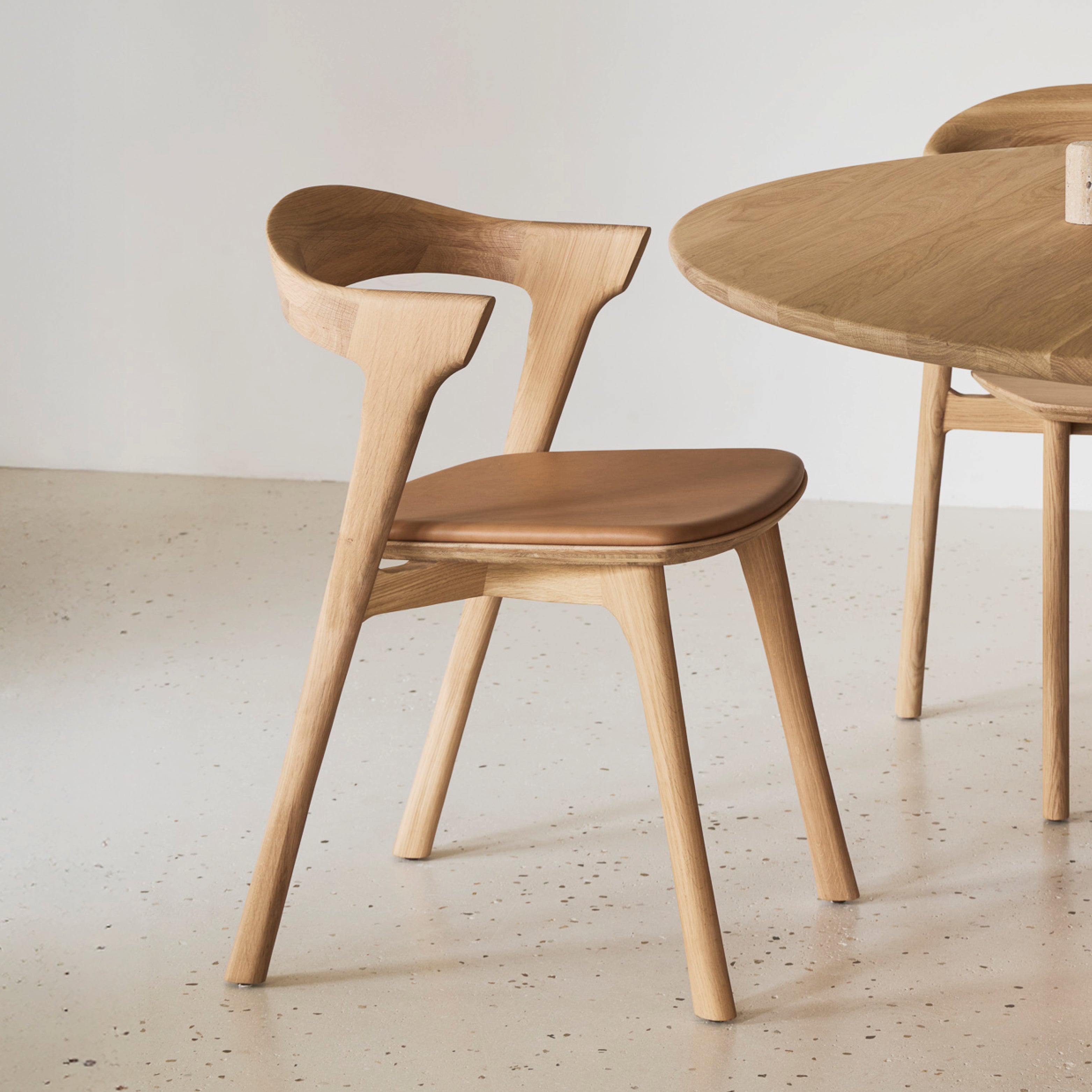 Ethnicraft Bok Stuhl aus massiver Eiche mit geschwungener Rückenlehne und Sitzkissen aus cognacfarbenem Leder, freistehend im Tageslicht auf heller Oberfläche.