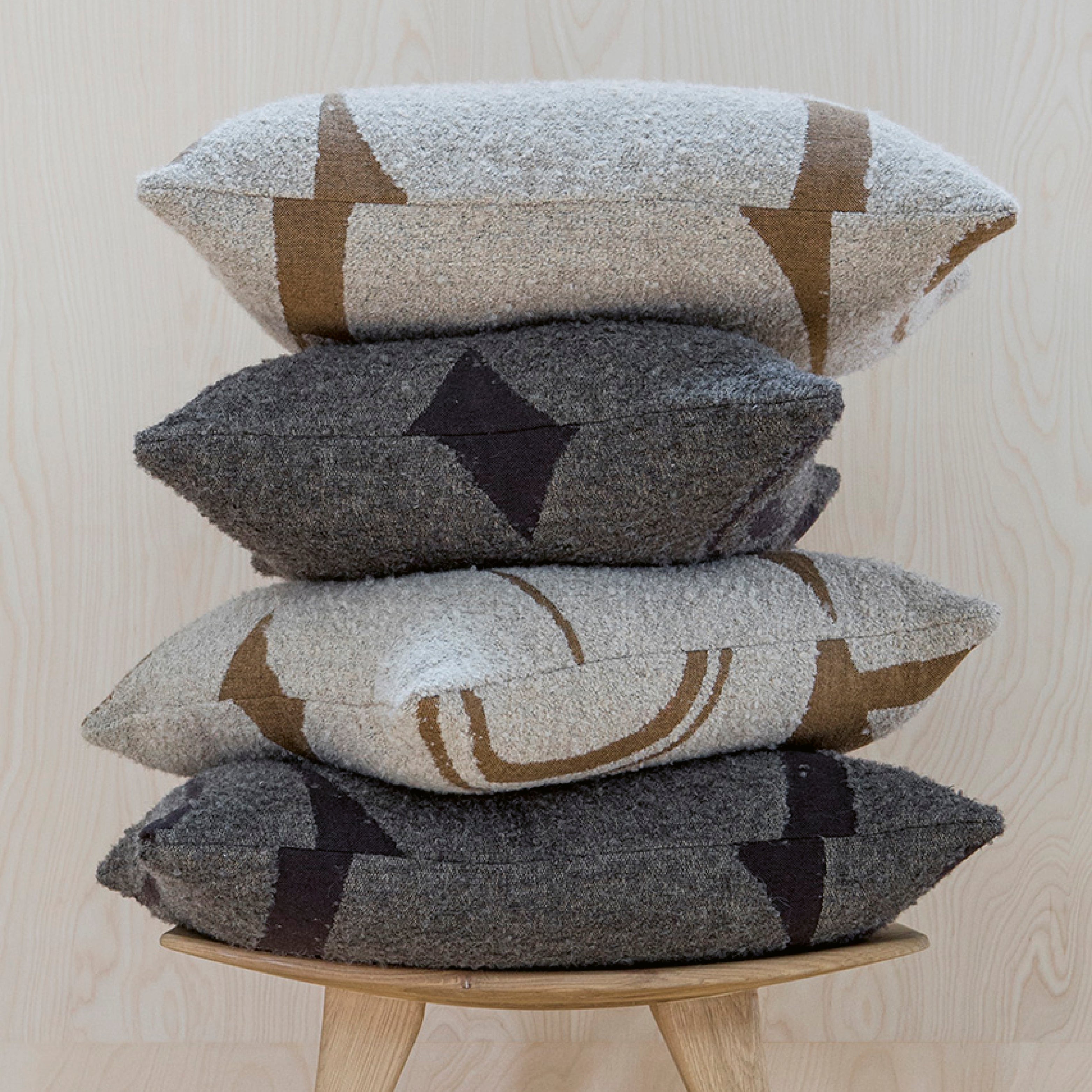 Vier grafisch gemusterte Ethnicraft-Kissen in Naturtönen, übereinandergestapelt auf einem Hocker vor hellem Holzhintergrund – minimalistisches, weiches Wohnstatement.