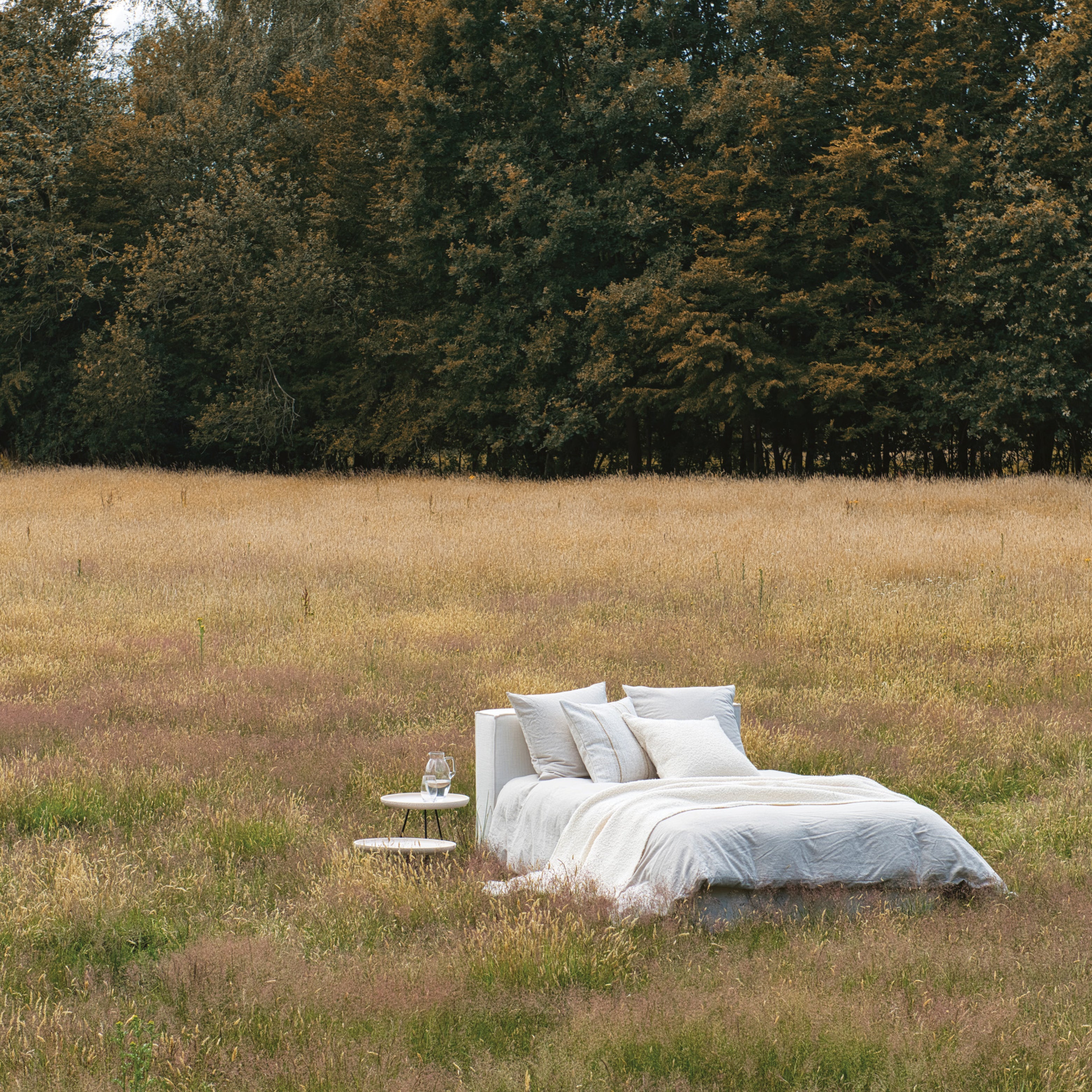 Natürliches Schlafzimmer-Setting: Bett mit heller Bettwäsche mitten auf einer Wiese vor einem Wald – Natural Homestorys Stimmung zwischen Natur und Rückzugsort.