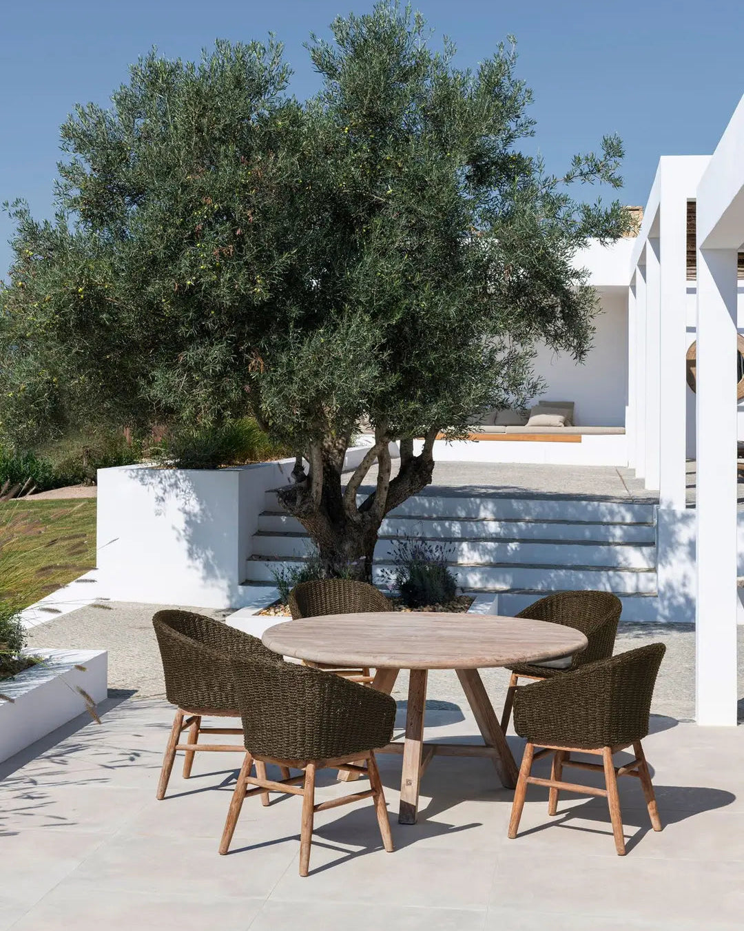 Esstisch ANTON Ø120 cm von Gommaire in Patio-Szene – runder Teaktisch in stilvoller Outdoor-Umgebung mit Natursteinboden und Gartenaccessoires