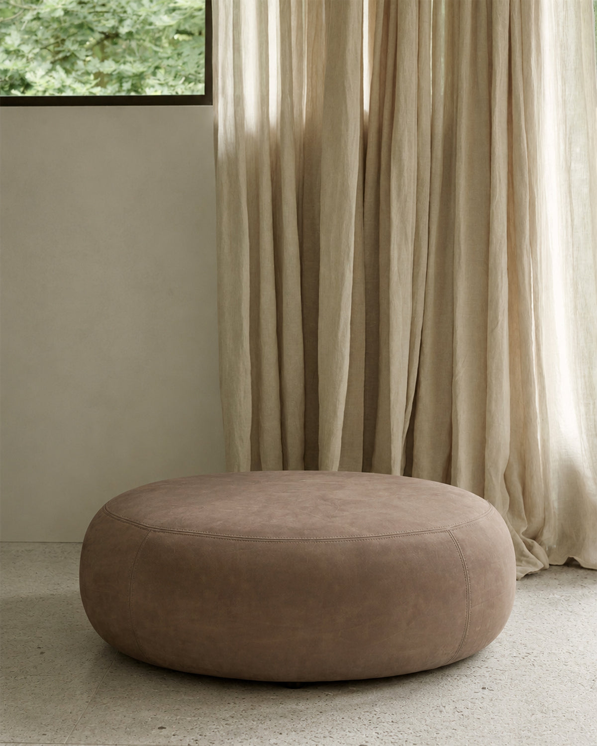 Ethnicraft ORB Sitzhocker in Fawn Nubukleder als gepolsterter Fußhocker vor einem Loungesessel in einem gemütlichen Wohnbereich.