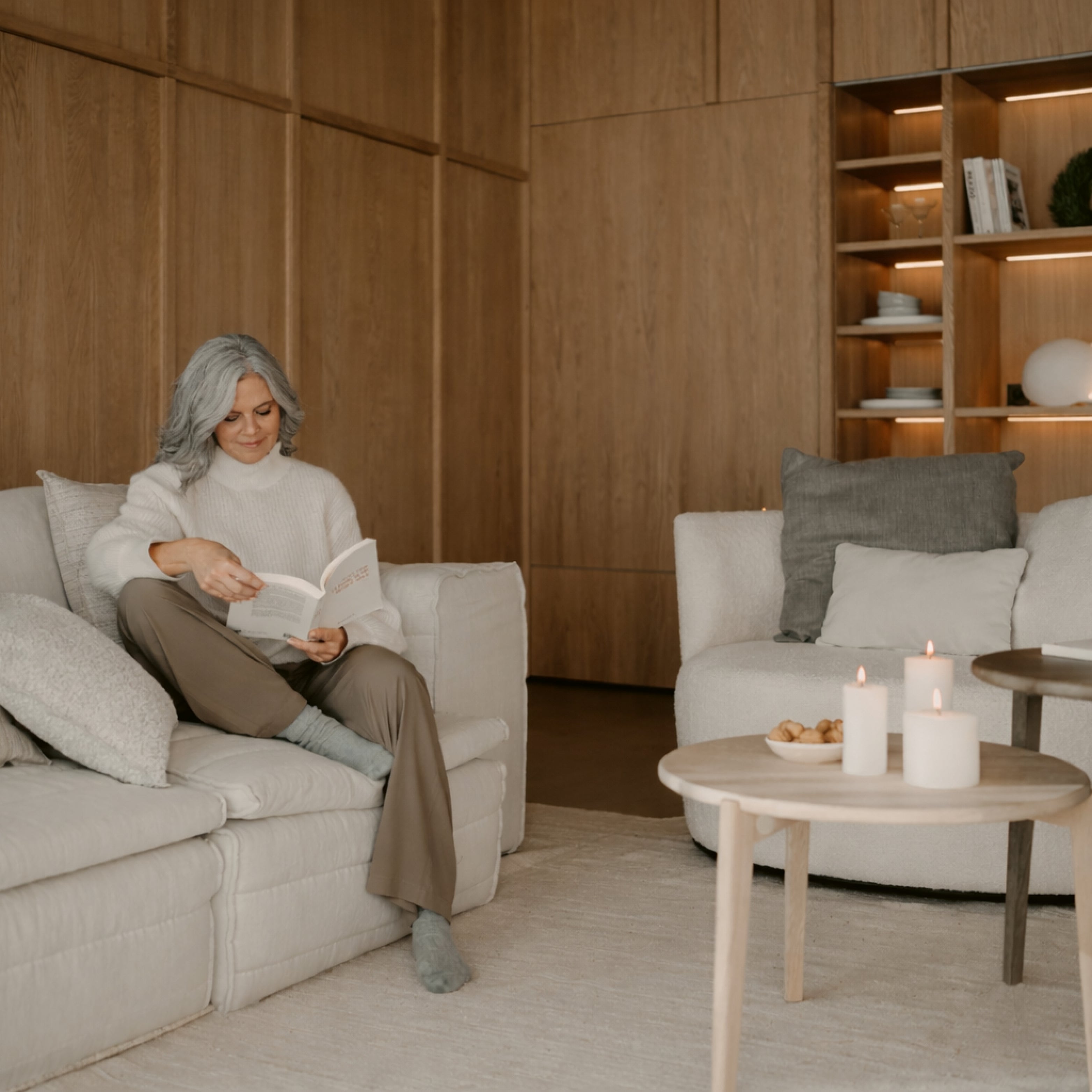 Frau mit grauen Haaren liest entspannt auf einem modernen, cremefarbenen Sofa in einem stilvoll eingerichteten Wohnraum mit Holzverkleidung und skandinavischem Flair.