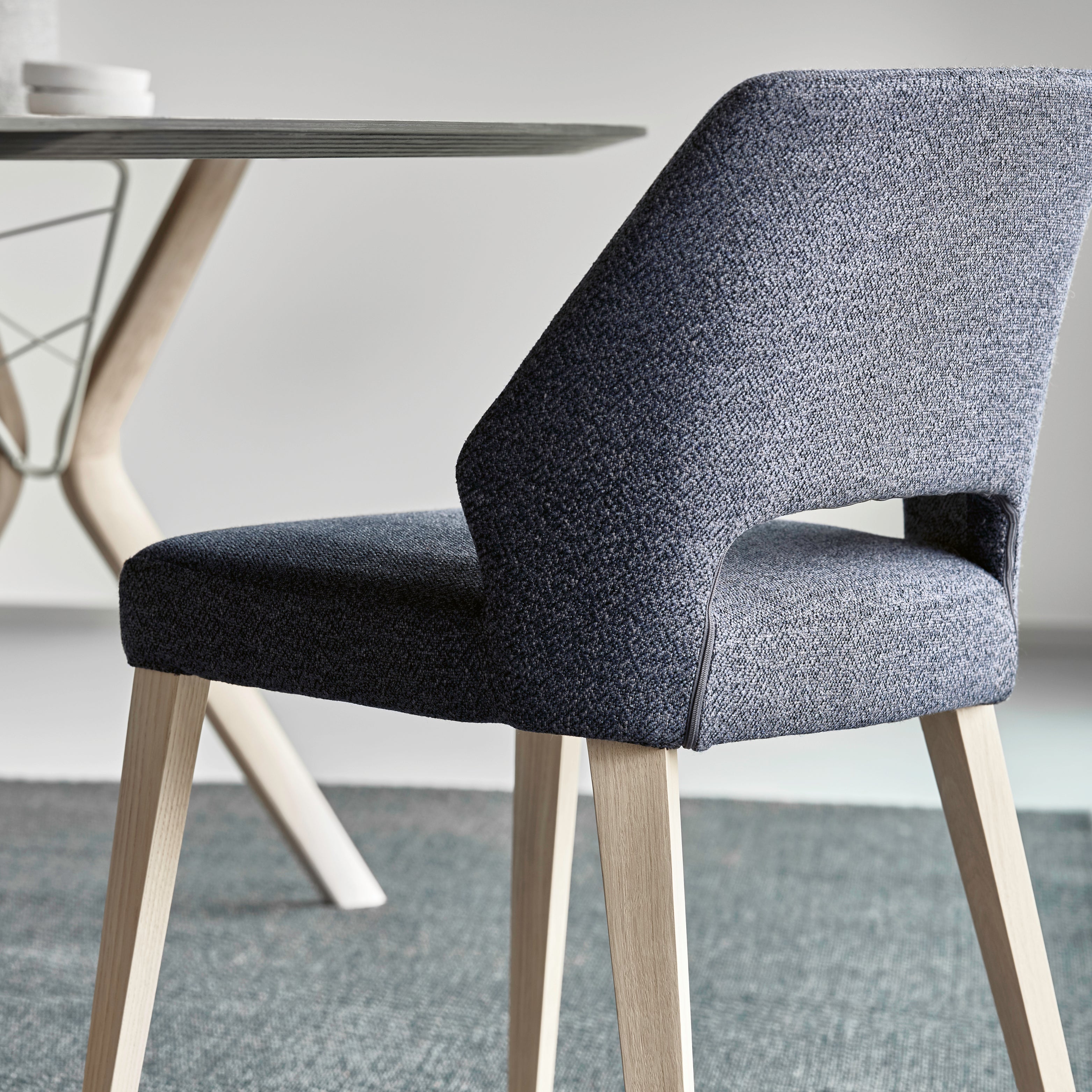 Mobitec Lena Stuhl mit anthrazitfarbenem Stoffbezug, charakteristischer Rückenaussparung und hellem Holzgestell – modernes Sitzdesign vor rundem Esstisch mit skulpturalem Untergestell.