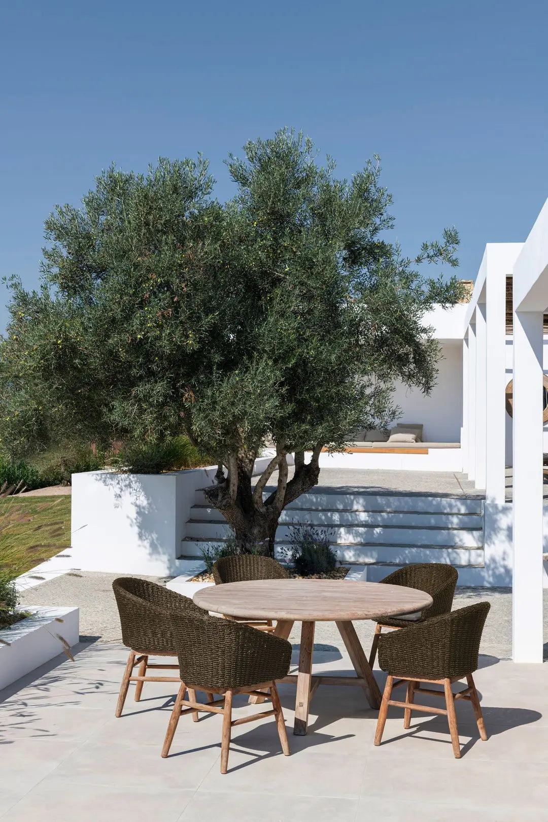 Esstisch ANTON Ø120 cm von Gommaire in Patio-Szene – runder Teaktisch in stilvoller Outdoor-Umgebung mit Natursteinboden und Gartenaccessoires