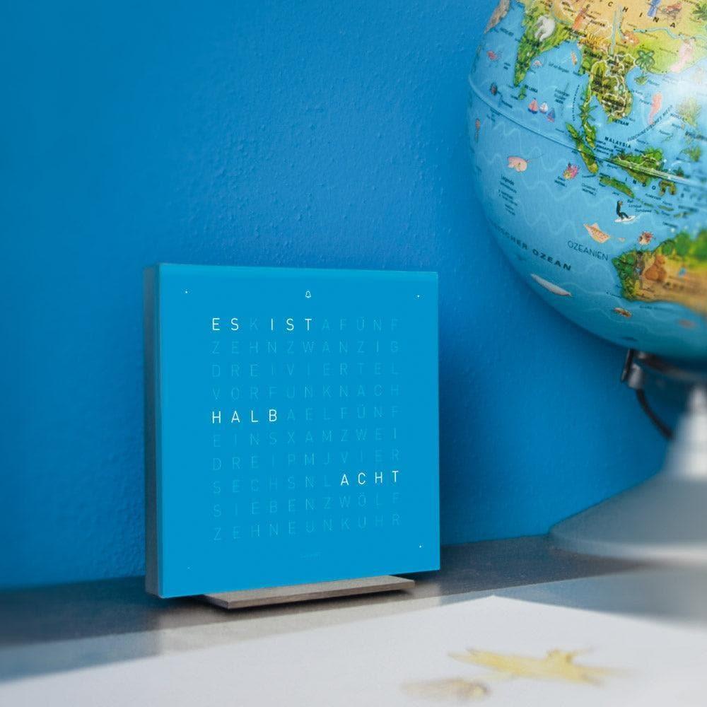 QLOCKTWO EARTH 13.5 Blue Candy – moderne Design-Tischuhr mit typografischer Zeitanzeige in kräftigem Blau, stilvoll inszeniert auf Möbelstück im Interior-Setting